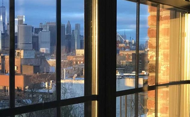 Interior view of floor-to-ceiling windows with Manhattan skyline visible, including Empire State Building and East River, black window frames with yellow interior light
