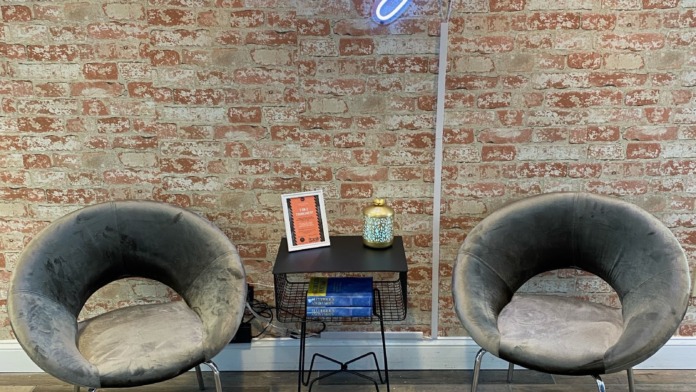 Interior lounge with two curved modern gray chairs, small side table, exposed brick wall, neon signage, and wood flooring