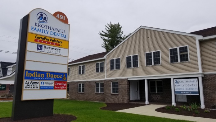 Exterior view of brick and vinyl-sided building with directory sign listing multiple business names and landscaped grounds