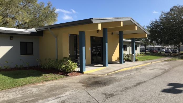 Exterior view of main entrance building with yellow and gray colored walls, covered portico, and parking area visible