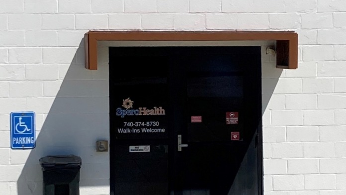 Exterior entrance showing black door with Spero Health signage, white brick wall, handicap parking marking, and welcome information