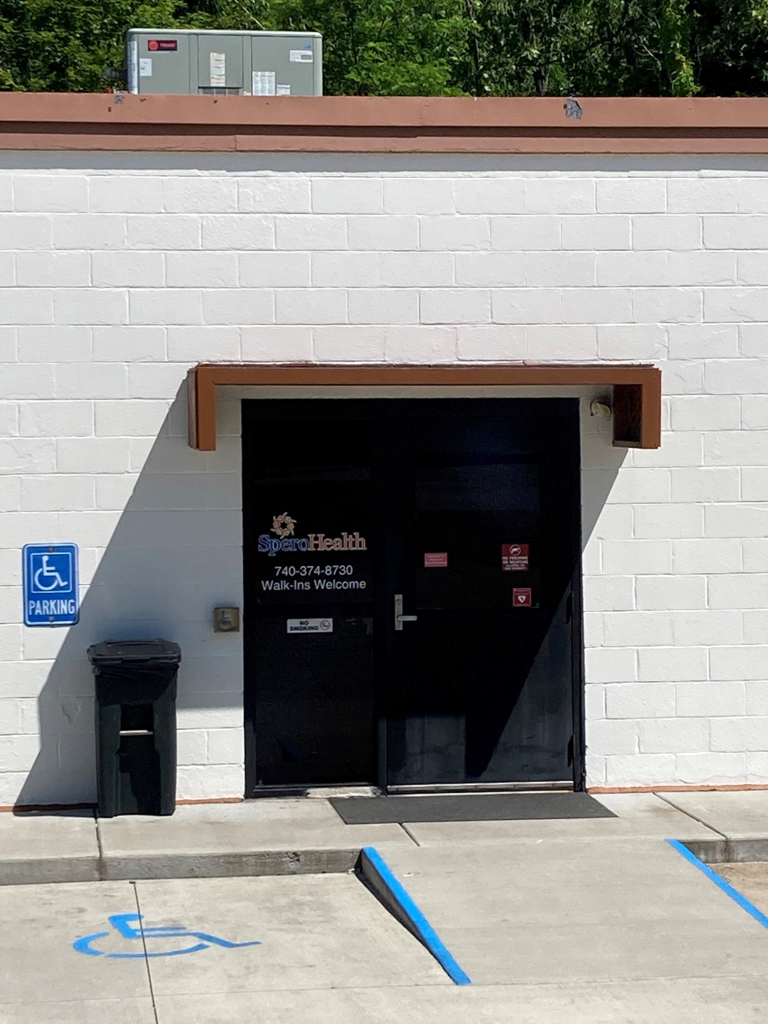 Exterior entrance showing black door with Spero Health signage, white brick wall, handicap parking marking, and welcome information