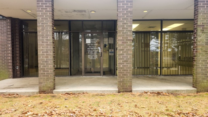 Exterior brick facade with glass entrance doors, vertical blinds, concrete patio, structural columns, and minimal landscaping