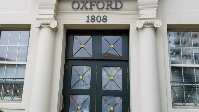 Exterior of neoclassical building entrance featuring white columns, black double doors marked Oxford 1808, and wrought iron railings.