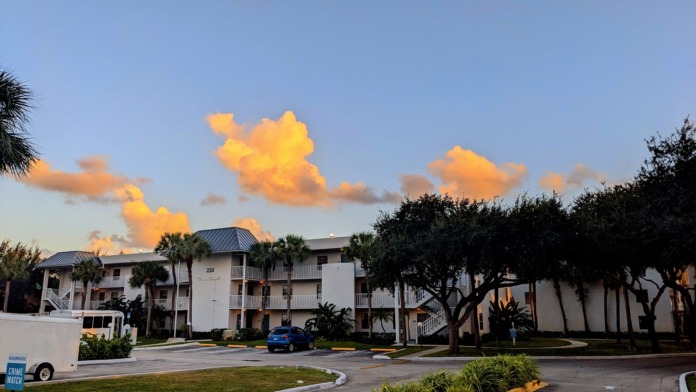 Exterior view of multi-story residential building with white facade, metal railings, palm trees, and parking area at sunset