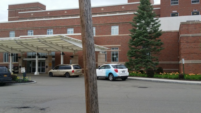 Exterior view of medical facility entrance with brick construction, covered portico, parked vehicles, and landscaping.