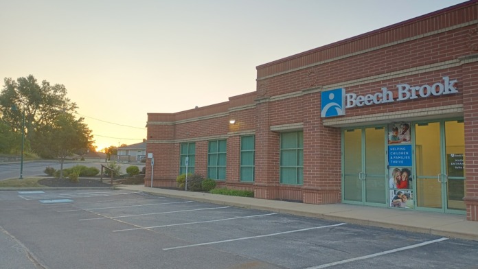Exterior view of Beech Brook treatment center showing red brick building facade, main entrance with turquoise doors, Beech Brook logo signage, and parking area.