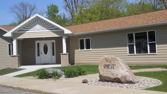 Exterior of treatment facility showing main entrance with portico, tan siding, landscaped grounds with engraved facility stone