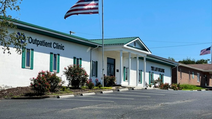 Exterior of VA outpatient clinic showing main entrance with columns, green trim, landscaping, and parking area.