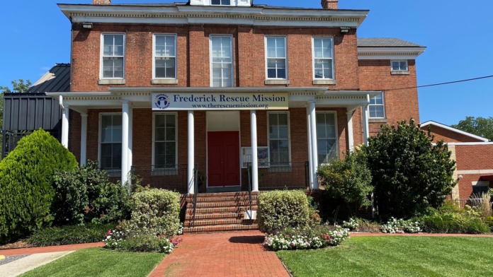 Exterior front of historic brick building with white columns, covered porch, dormer windows, brick walkway, and manicured landscaping