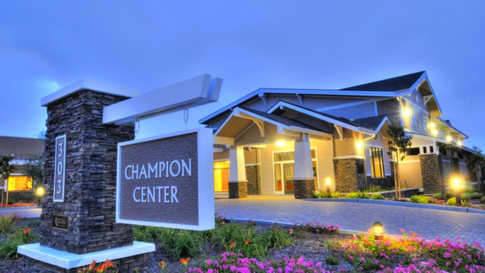 Exterior view of Champion Center showing illuminated main entrance, stone signage, manicured landscaping with colorful flowers, and paved driveway at dusk