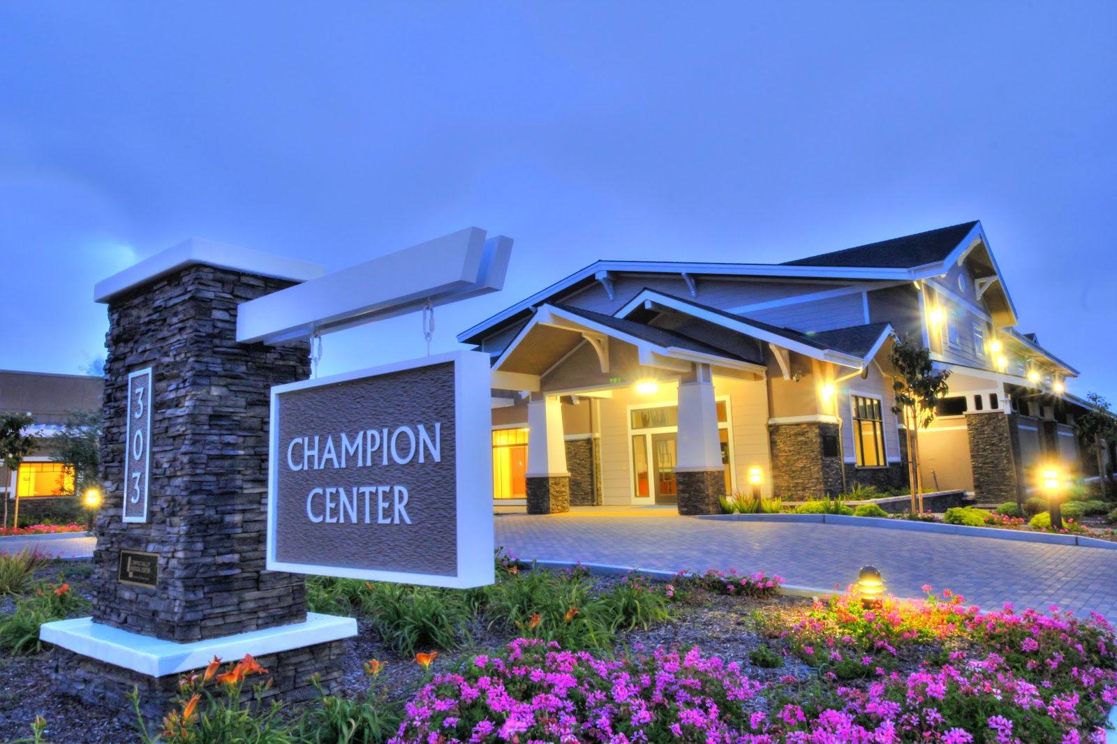 Exterior view of Champion Center showing illuminated main entrance, stone signage, manicured landscaping with colorful flowers, and paved driveway at dusk