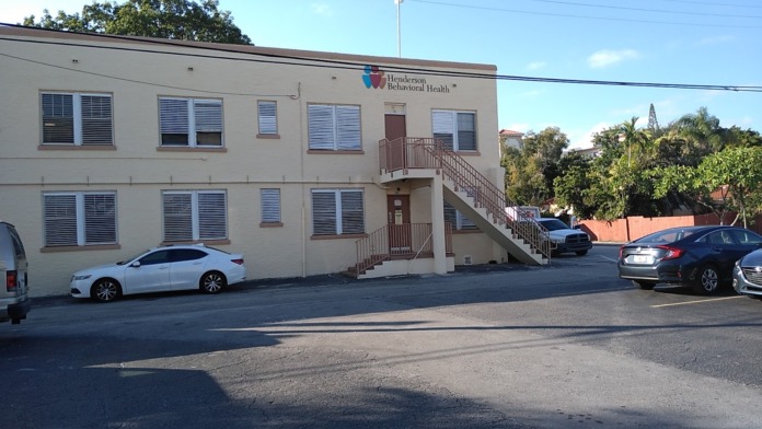 Exterior view of Henderson Behavioral Health facility showing two-story building facade, main entrance with stairs, parking area with vehicles, and signage