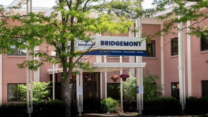 Exterior of brick facility building with white columns, porch, pink and tan facade, Bridgemont signage, mature tree and landscaping.