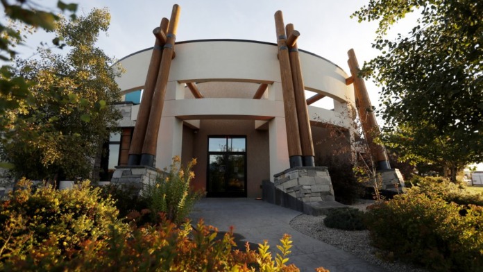 Exterior front entrance featuring rounded modern building with tall wooden architectural supports, landscaping, and mature trees.