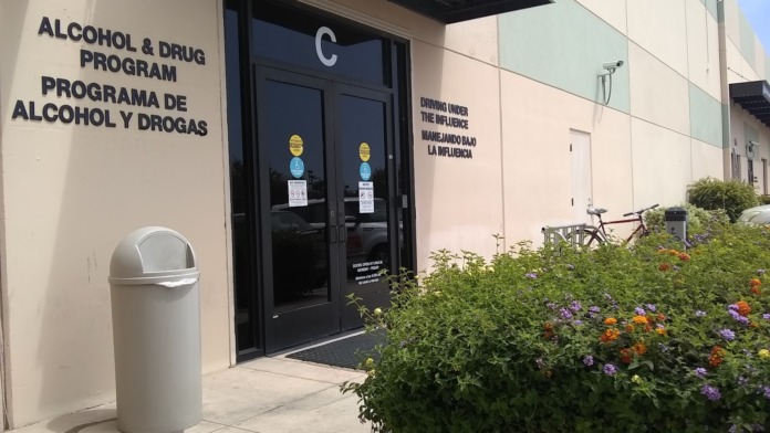 Exterior entrance showing Alcohol & Drug Program bilingual signage, black doors marked C, trash bin, and landscaped flower beds