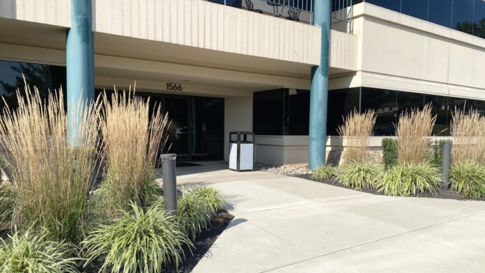Building exterior ground-level entrance showing concrete columns, ornamental grasses landscaping, sidewalk, address number 1566, and modern structure.