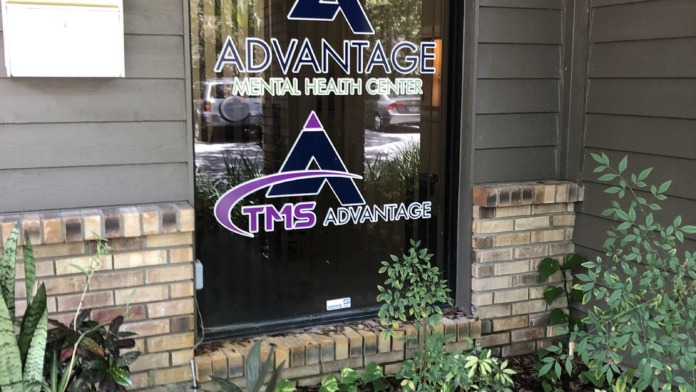 Exterior entrance door displaying facility logos with brick exterior, landscaped grounds, and ornamental plants