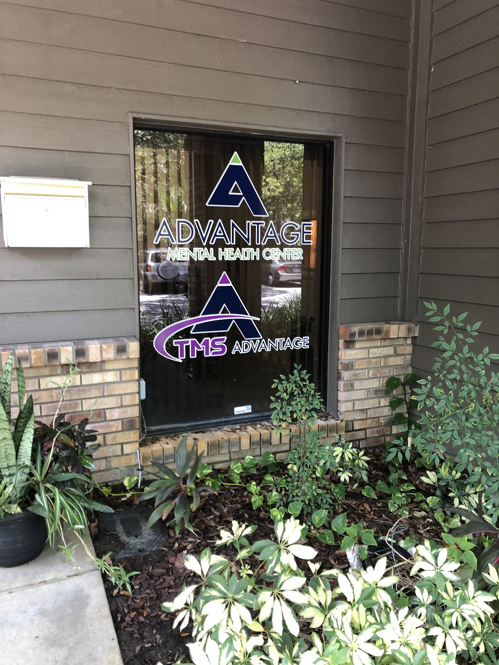 Exterior entrance door displaying facility logos with brick exterior, landscaped grounds, and ornamental plants
