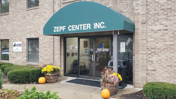 Exterior front entrance of Zepf Center Inc. showing teal awning, glass doors, decorative planters with fall flowers, and facility signage