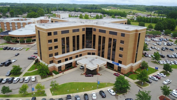 Exterior aerial view of modern multi-story medical facility with tan brick exterior, white rooflines, and expansive parking areas with landscaping.