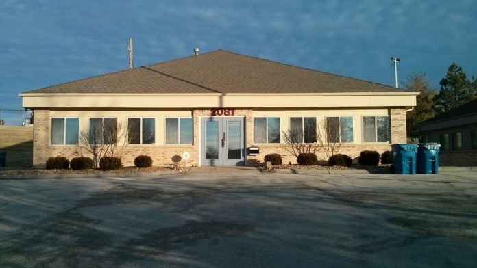 Exterior view of single-story brick treatment center building with peaked roof, multiple windows, landscaping, and parking area.