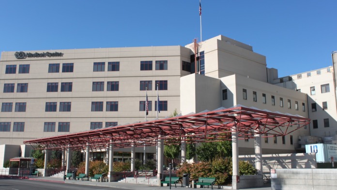Exterior front view of medical facility with tan colored building, red metal canopy entrance structure, flagpoles, and clear sky