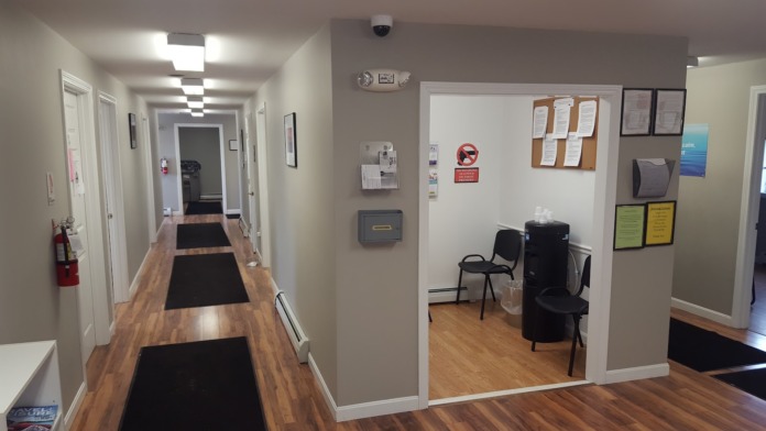 Interior hallway showing multiple white doors to patient rooms, wooden flooring, black runner mats, and facility signage