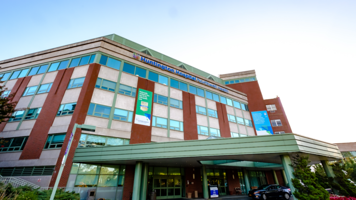 Exterior view of modern multi-story hospital building with brick and glass facade, covered main entrance canopy with green trim, glass doors, and parking area.