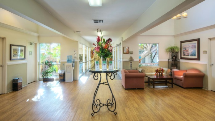 Interior spacious lobby with wooden flooring, sitting area with upholstered furniture, large central floral arrangement, and windows