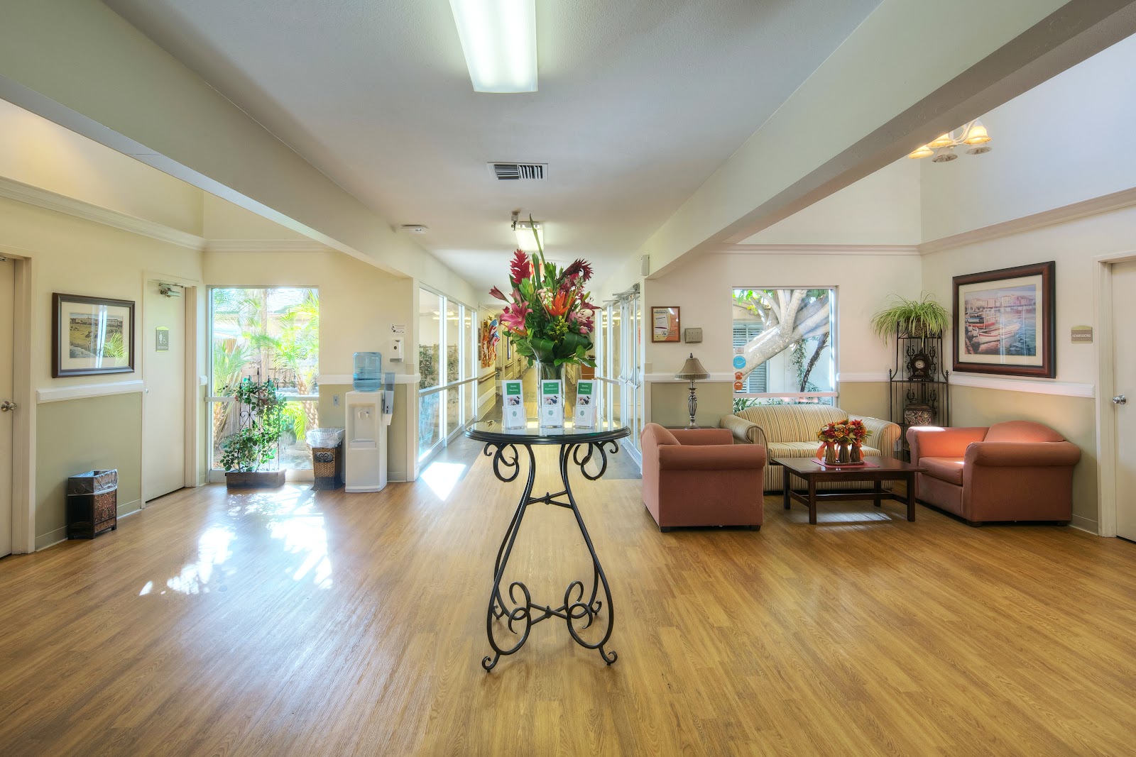 Interior spacious lobby with wooden flooring, sitting area with upholstered furniture, large central floral arrangement, and windows