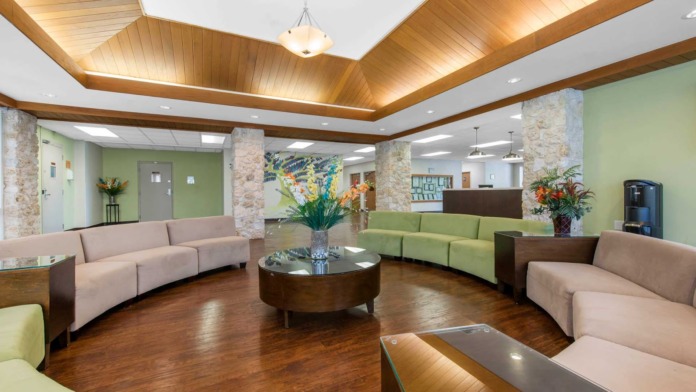 Interior lobby with exposed beam ceiling, green accent walls, curved seating arrangements in beige and green, reception desk, and stone pillars