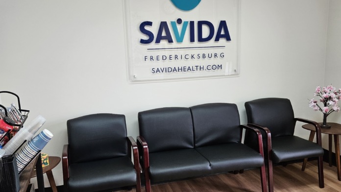 Interior reception waiting area with black leather seating, SaVida Health Fredericksburg logo, wooden flooring, and decorative flowers.