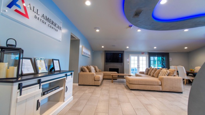 Interior reception lobby with All American Detox Center logo, comfortable seating, and modern blue ceiling design elements.