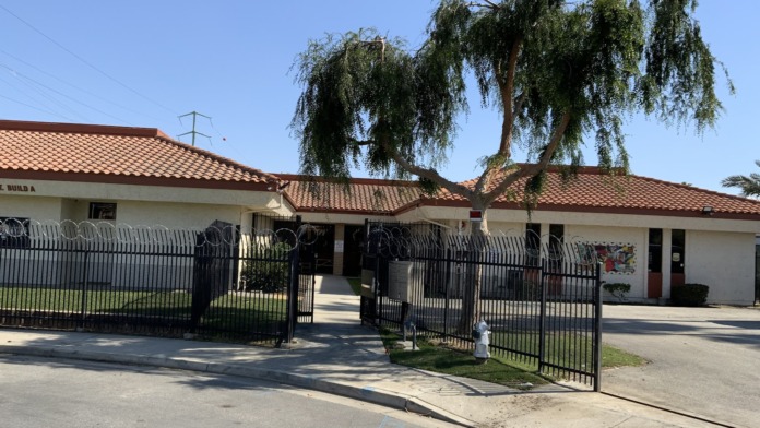 Street-facing exterior showing entrance gates, landscaping with weeping willow tree, facility building and parking area