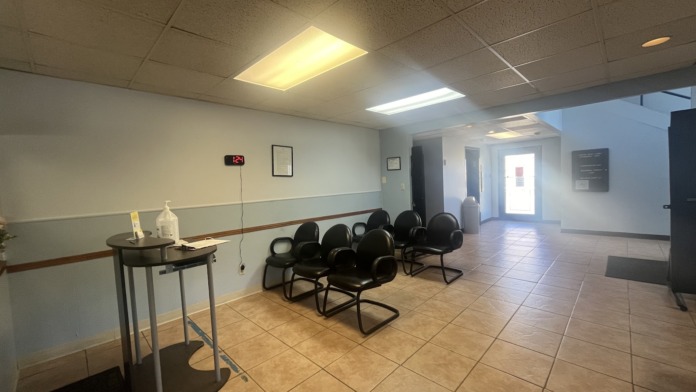 Interior waiting room with black chairs, tan tile flooring, fluorescent ceiling lights, and entry corridor with bright doorway.