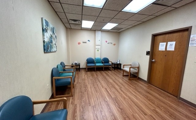 Interior waiting room featuring upholstered chairs, wood-paneled dividers, framed artwork, and drop ceiling lighting.