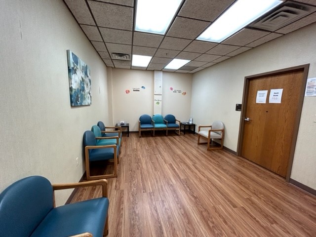 Interior waiting room featuring upholstered chairs, wood-paneled dividers, framed artwork, and drop ceiling lighting.