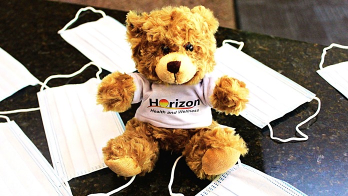 Brown teddy bear mascot wearing Horizon Health and Wellness branded shirt surrounded by medical masks on surface