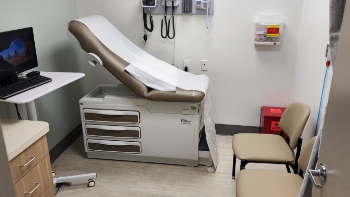 Interior clinical exam room featuring adjustable medical examination chair, diagnostic equipment on wall, desk with laptop, and chair seating