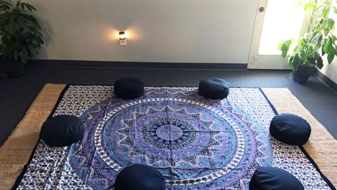 Interior meditation room with mandala rug, arranged black cushions in circle, potted plants, and natural light from window
