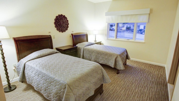 Interior patient room with two hospital beds with quilted bedding, wooden headboards, individual nightstands, windows, and private bathroom