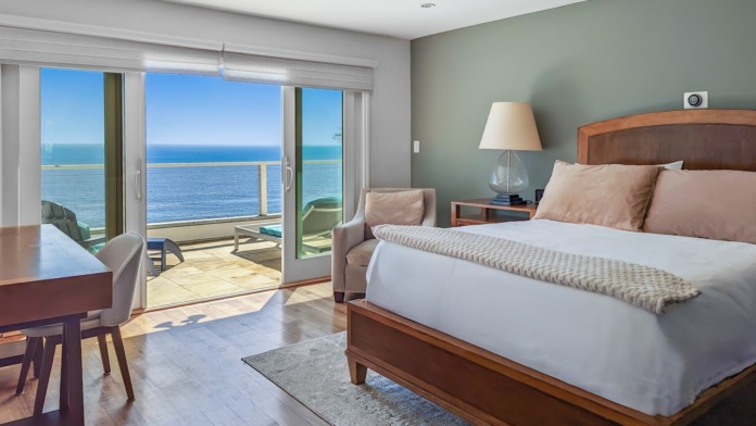 Interior patient bedroom with ocean-facing balcony, queen bed, desk, nightstands, and panoramic sea views through sliding doors
