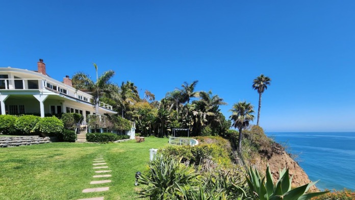 Exterior view of two-story beachfront residence with white columns, terrace, green lawn, and Pacific Ocean backdrop