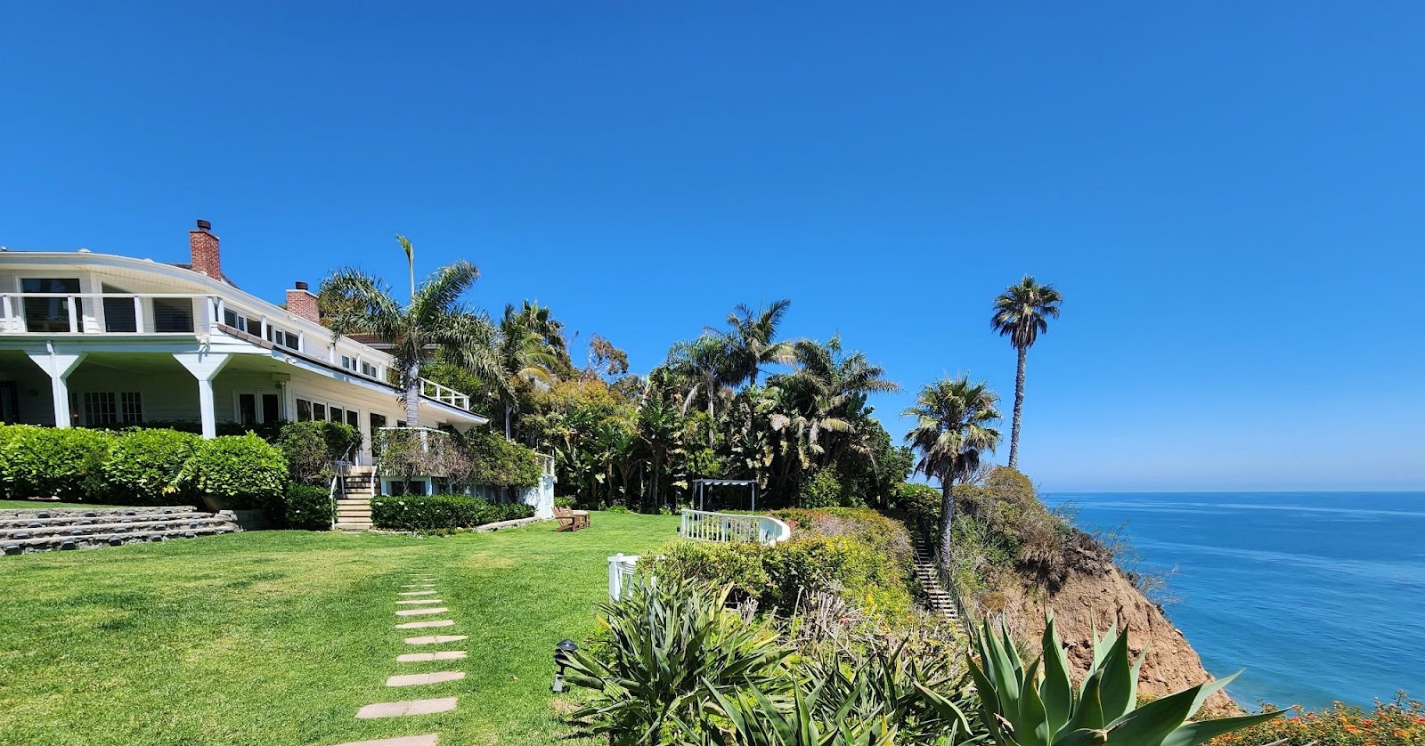 Exterior view of two-story beachfront residence with white columns, terrace, green lawn, and Pacific Ocean backdrop