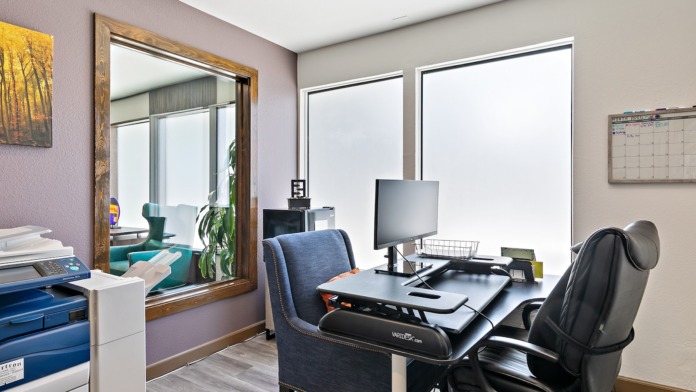 Interior office area with desk, computer monitor, ergonomic chair, mirror with wooden frame, and large bright windows
