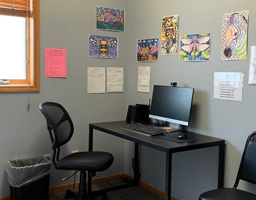 Interior office with gray walls, black desk with computer monitor and laptop, office chair, and colorful artwork