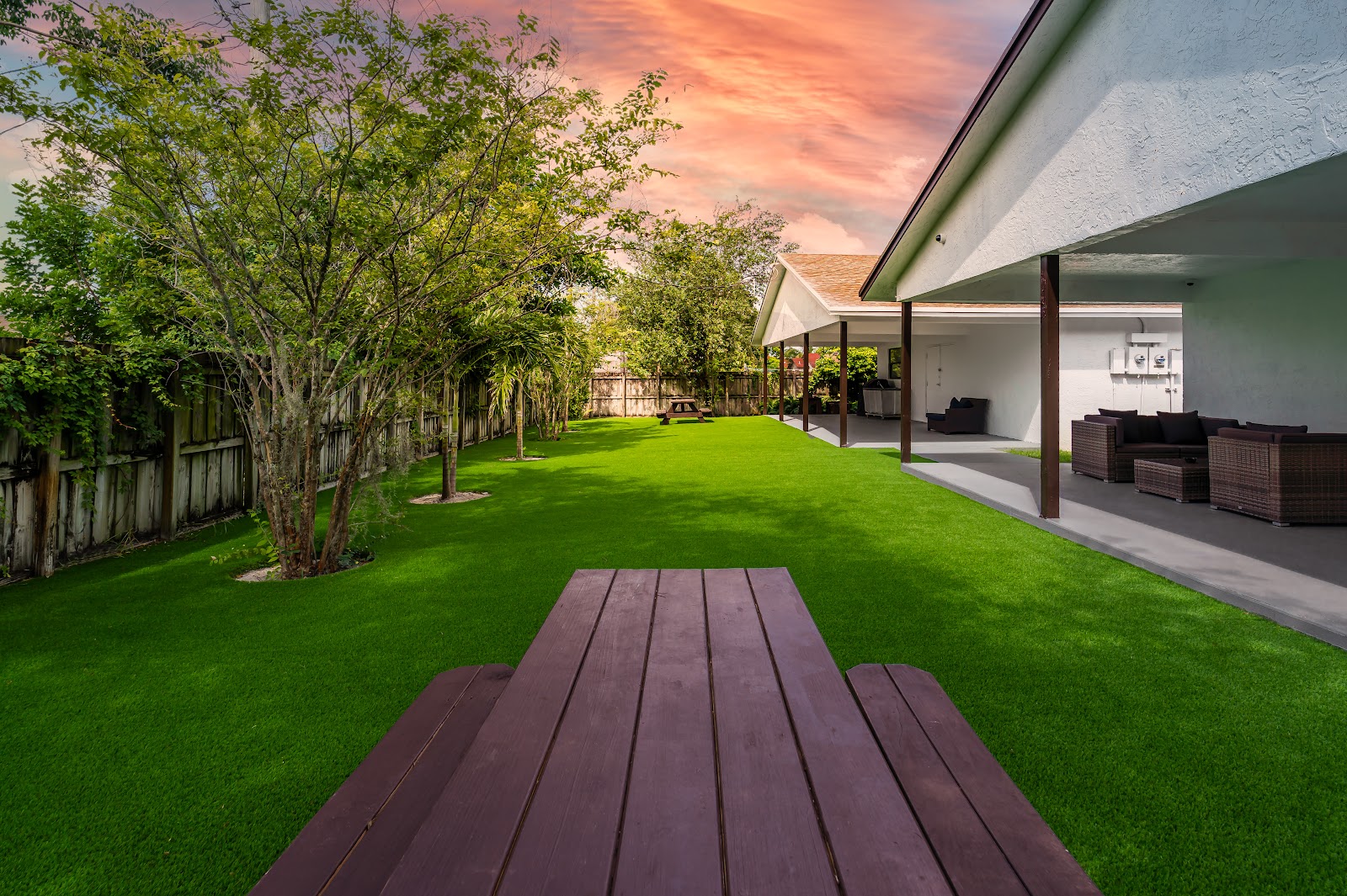 Exterior outdoor area with manicured lawn, mature trees, covered patio seating, and rattan furniture with sunset lighting