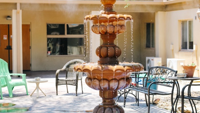 Exterior patio with tiered wooden fountain feature, lounge seating, fire pit, building with windows, tree shade coverage
