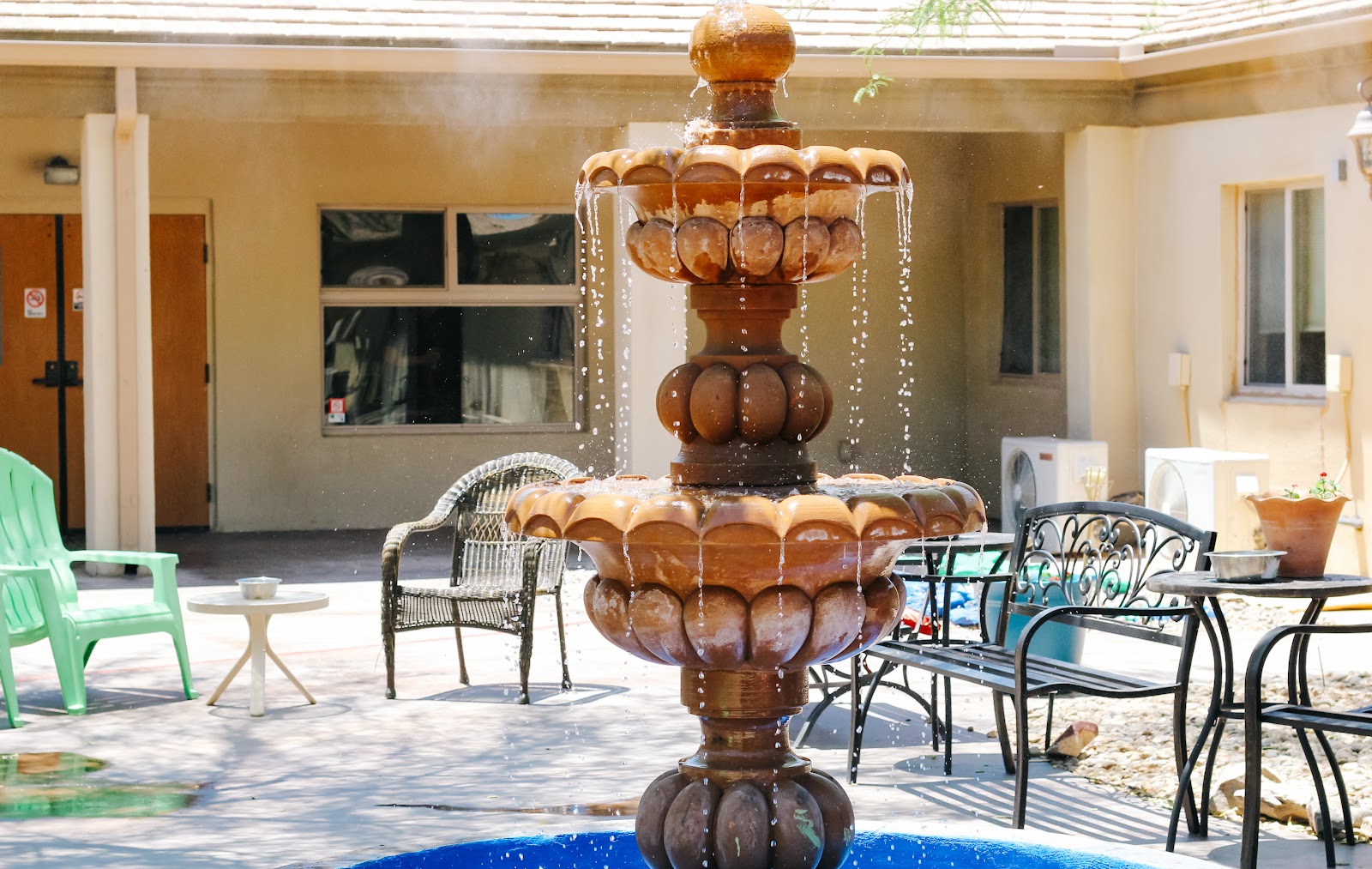 Exterior patio with tiered wooden fountain feature, lounge seating, fire pit, building with windows, tree shade coverage